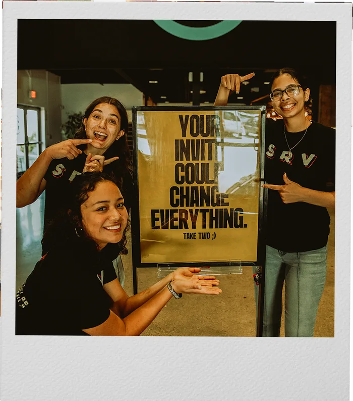 Volunteers pointing at a sign that reads: your invite could change everything